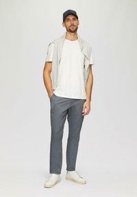 Grijze katoenbroek met een wit T-shirt en een lichtgrijze trui over de schouders. Witte sneakers en een marineblauwe pet maken de outfit af.
