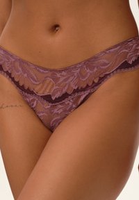 Lace thong in mauve with floral patterns, featuring sheer panels and a smooth waistband. Soft texture with intricate detailing.