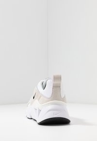 Nike-tennare som har en blanding av hvit og beige materiale, en polstret h æl, teksturert semsket skinn og en fremtredende svart logo på siden.