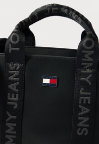 Mustast tekstureeritud sünteetilisest materjalist kandekott, millel on logo märgistus ja kootud rihm tekstiga 'TOMMY JEANS'.
