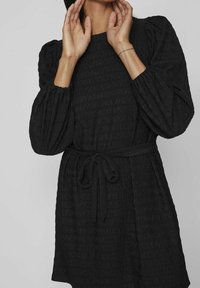 Una persona que lleva un vestido negro texturizado con mangas abullonadas y una cintura atada posa con las manos cerca del cuello contra un fondo neutro.