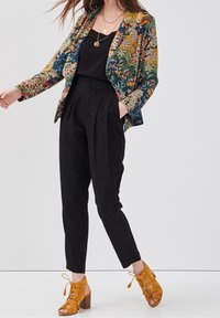 Woman wearing black high-waist trousers, black top, floral printed blazer, layered gold necklaces, and mustard lace-up heeled sandals.