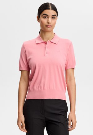 Donna con capelli scuri raccolti in una coda bassa, indossa una polo rosa a maniche corte e pantaloni neri, in piedi davanti a uno sfondo chiaro e uniforme.