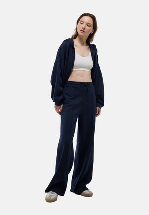Haut à capuche oversize bleu marine et pantalon de survêtement large en tissu doux, associé à un haut court blanc. Comprend une taille élastiquée avec cordon et des pantoufles décontractées.