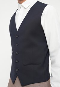 Colete azul-marinho feito de tecido suave, com um decote em V profundo, seis botões e um único bolso lateral. Combinado com uma camisa branca de colarinho.
