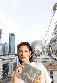 Silver metallic clutch bag with a textured surface and an infinity symbol detail. The bag is held against a cityscape background.