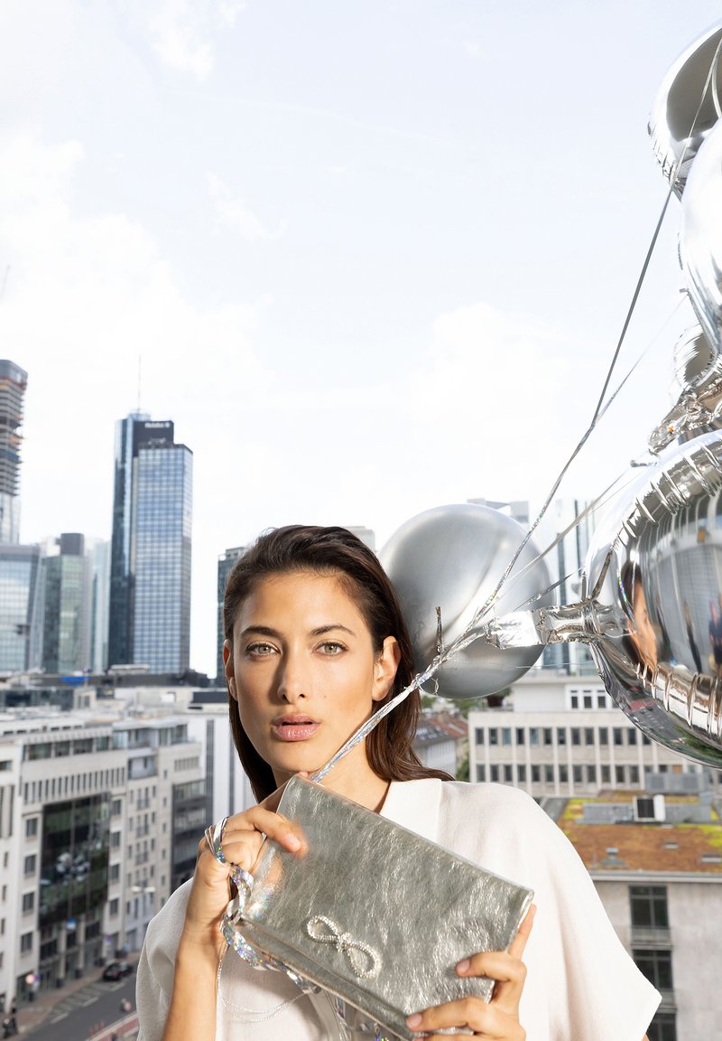 Silver metallic clutch bag with a textured surface and an infinity symbol detail. The bag is held against a cityscape background.