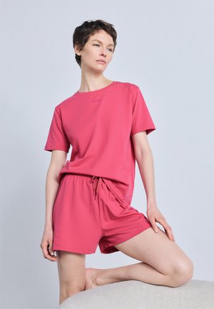 Kobieta z krótkimi włosami ubrana w komplet koralowo-różowy: t-shirt i szorty, pozująca z jedną kolanem opartym na zakrzywionej powierzchni.