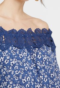 Blå topp med avskuren axel som har ett blommönster med vita blommor, detaljerad med en mörkblå spetskant runt halsringningen.