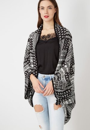 Mujer vistiendo una blusa negra con encaje, jeans claros desgastados y un chal suelto de estampado en blanco y negro contra un fondo liso.