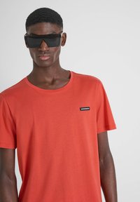 Crvena pamučna majica s okruglim izrezom i kratkim rukavima, s malim crnim oznakom brenda na lijevoj strani prsnog predjela. Crne oversized sunčane naočale.