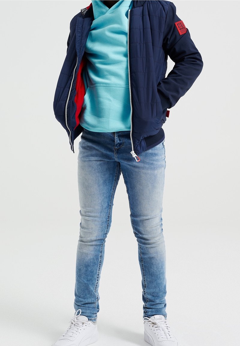 Námořnický prošívaný kabát s červenou podšívkou, tyrkysová mikina s kapucí, světle modré džíny a bílé tenisky. Zapínání na zip vpředu a detail s logem.