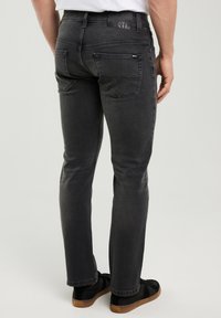 Sötétszürke egyenes szárú farmernadrágot és fekete sportcipőt viselő férfi, háttal, egyszínű háttér előtt.