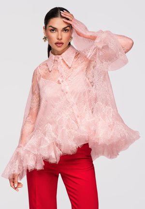 Kobieta w przezroczystej różowej koronkowej bluzce z falbankami na rękawach i kołnierzyku, w jasnoczerwonych spodniach, pozująca z ręką na czole.