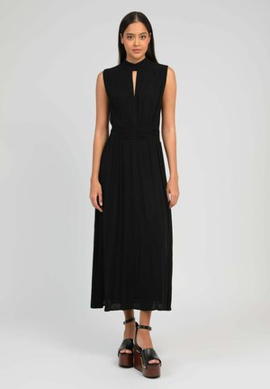 Femme portant une robe midi noire sans manches avec un décolleté en forme de goutte et une taille froncée, associée à des sandales plateformes noires.