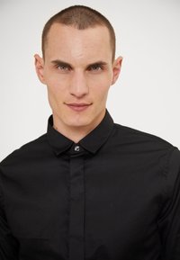 Camisa negra de botones, con textura suave, corte ajustado y tapeta oculta. Presenta un pequeño logo en el lado izquierdo del pecho.