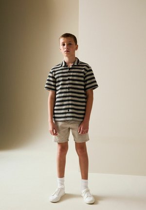 Short-sleeved, collared shirt with black and white zigzag pattern, paired with beige shorts and white sneakers. Background is a light neutral color.