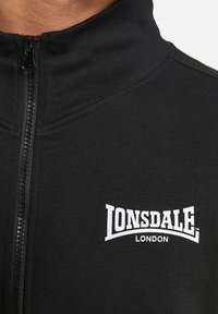Chaqueta negra con cremallera hecha de tela acanalada, con un alto cuello y un logo blanco de 'Lonsdale London' en el pecho.