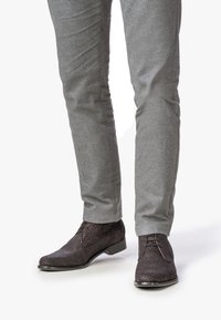 Zapatos de cuero trenzado marrón oscuro con un tacón bajo, con cordones y una textura intrincada. Combinados con pantalones grises.