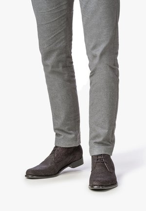Dunkelbraune, geflochtene Lederschuhe mit niedrigem Absatz, versehen mit Schnürsenkeln und einem kunstvollen Muster. Kombiniert mit grauen Hosen.