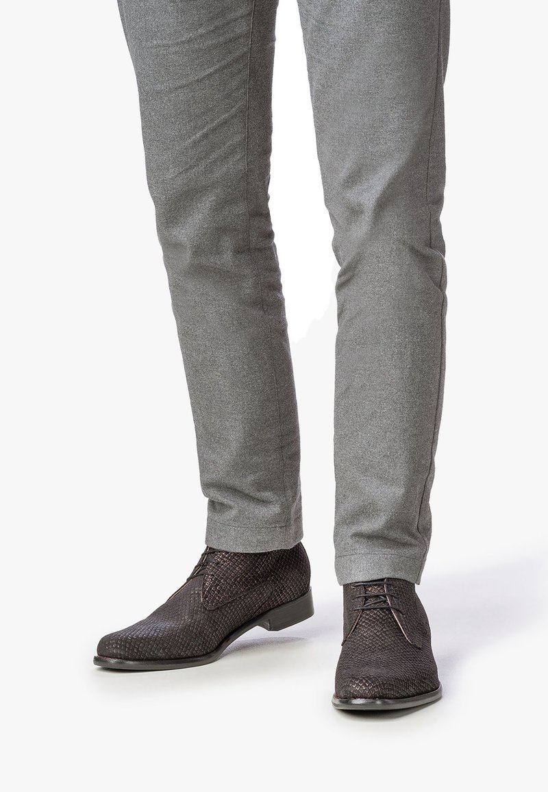 Zapatos de cuero trenzado marrón oscuro con un tacón bajo, con cordones y una textura intrincada. Combinados con pantalones grises.