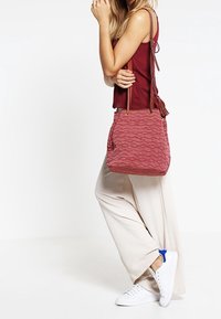 Rød tekstureret spandtaske med diagonale bølgemønstre, med to skulderremme og lynlåslukning. Båret over et lysefarvet outfit.