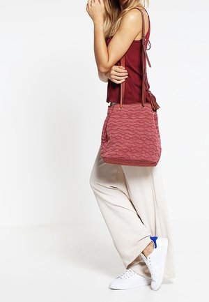 Punainen, kuvioitu olkalaukku, jossa on vinottain aaltoilevat kuviot, kaksi olkahihnaa ja vetoketjusuljin. Laukku on pukeutunut vaalean värisen asun päälle.