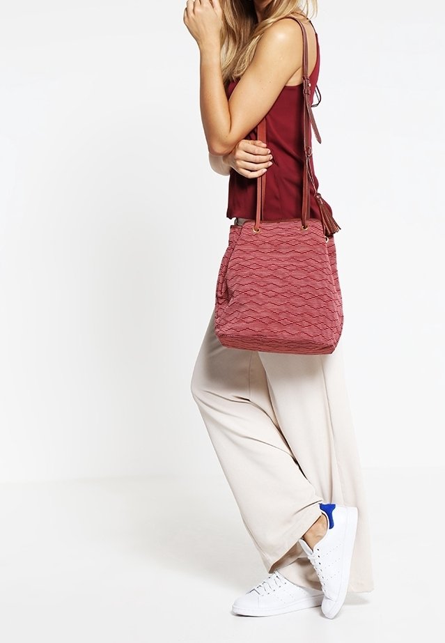 Rød tekstureret spandtaske med diagonale bølgemønstre, med to skulderremme og lynlåslukning. Båret over et lysefarvet outfit.