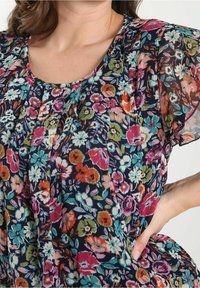 Blomstret bluse med en mørk marineblå bund og blomster i pink, grøn og orange. Gennemsigtig tekstur, plisseret design og korte flagermusærmer.