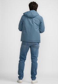 Chaqueta de plumas azul con capucha, que presenta una textura suave y puños elásticos. Combinada con jeans azul claro y zapatos blancos.