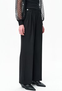 Zwarte broek met wijde pijpen, voorzien van een platte voorkant, een enkele knoop als accent en een gladde stoftextuur. Draagt bijpassende puntige zwarte schoenen.