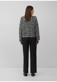 Vrouw die met haar rug naar de camera staat, gekleed in een grijze sweater met luipaardprint, zwarte wijde pantalon en zwarte schoenen, tegen een effen grijze achtergrond.