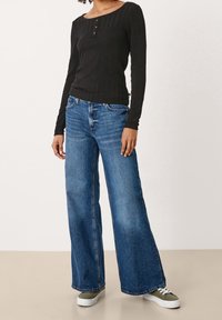 Schwarzes, geripptes Langarmoberteil mit einem Rundhalsausschnitt und Knopfdetails, kombiniert mit weit geschnittenen blauen Jeans und grünen Turnschuhen.