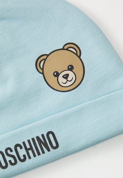 Šviesiai mėlynas audinys su šypseną rodančio meškiuko veido grafika ir ryškiu juodu "MOSCHINO" tekstu, atspausdintu ant rankovės.