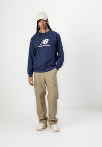 Misiņa krāsas džemperis ar baltu "New Balance" logo, bēšas krāsas pārsēju bikses un krējuma krāsas sporta apavi, apvienoti ar gaišas krāsas cepuri.