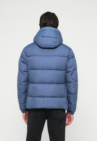 Sinine puhvjak Jacket, millel on eemaldatav kapuuts, horisontaalselt tikitud sektsioonid ja soonikust käised, kantud tumedate teksade peal.