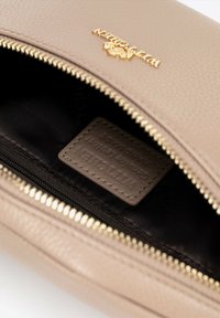 Beżowa skórzana saszetka z złotym detalem zasuwki. Wnętrze ma ciemne wykończenie i prostokątną etykietę z napisem "skóra naturalna."