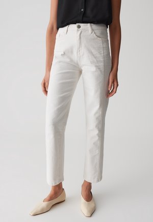 Vrouw met hoge taille witte spijkerbroek, zwart mouwloos topje en beige puntige ballerina's, staand tegen een effen grijze achtergrond.