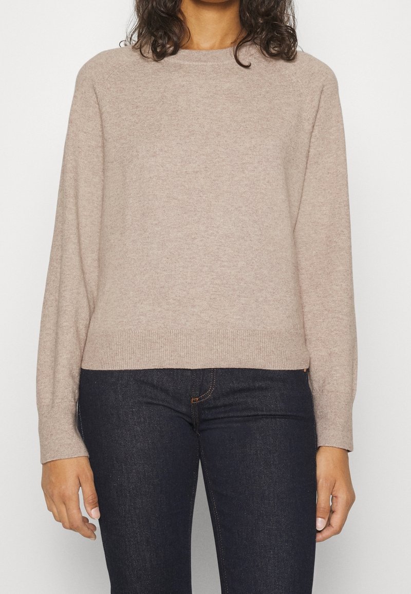 Femme portant un pull beige à manches longues et col rond, associé à un jean denim bleu foncé, visible de la poitrine aux hanches.