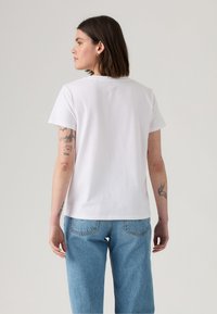 T-shirt en coton blanc à manches courtes, avec un col arrondi. Porté avec un jean décontracté bleu clair et des tatouages visibles sur les bras.