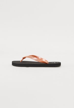 Chanclas negras con una amplia tira de cobre brillante que presenta un logo en relieve. Suela texturizada para agarre, diseño minimalista.