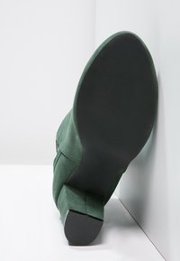 Zapato verde de ante con tacón alto mostrado desde un ángulo, que presenta una suela de goma negra sólida y un acabado texturizado en la parte superior.