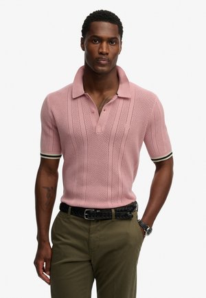 Różowa dzianinowa polo z teksturowanym wzorem, krótki rękaw i klasyczny kołnierzyk. Posiada czarno-białe akcenty na rękawach oraz luźny krój.