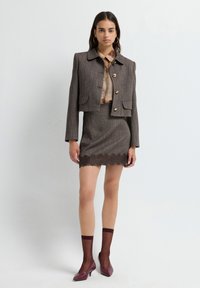 Grijze tweed cropped jas met gouden knoop, gecombineerd met een bijpassende rok met kantafwerking. Bourgondische puntige hakken en sheer sokken maken de look compleet.