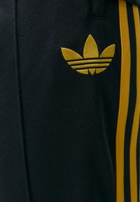 Melni sporta bikses ar dzeltenām svītrām un dzeltenu izšūtu logotipu ar ziedu motīvu kreisajā pusē. Strukturēta auduma.