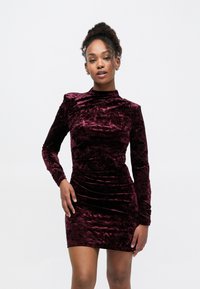 Robe en velours bordeaux à manches longues, col montant, et silhouette ajustée avec des détails froncés pour une texture supplémentaire.