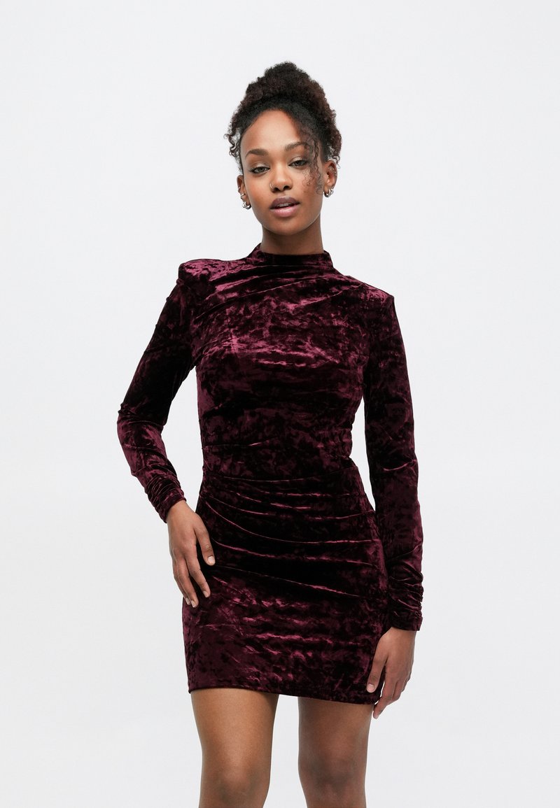Robe en velours bordeaux à manches longues, col montant, et silhouette ajustée avec des détails froncés pour une texture supplémentaire.