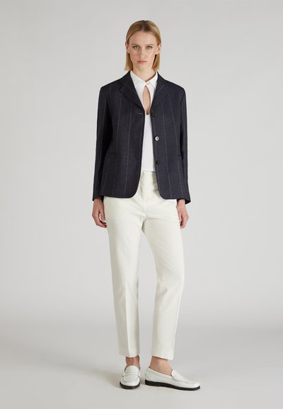Donna che indossa un blazer blu a righe, top bianco, pantaloni crema e mocassini bianchi, in piedi davanti a uno sfondo grigio semplice.