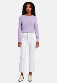 Pull en lilas texturé, jeans blancs évasés avec un ourlet effiloché et chaussures à enfiler crème. Coiffé de cheveux bouclés lâches et d'accessoires minimalistes.