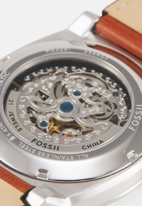 Boîtier de montre en acier inoxydable argenté avec un mouvement mécanique complexe visible. Bracelets en cuir marron. Détails notables en bleu et violet.